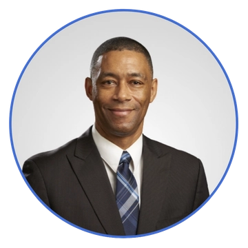 Professional headshot: smiling man in a dark suit and blue plaid tie, against a light gray background, circular frame.