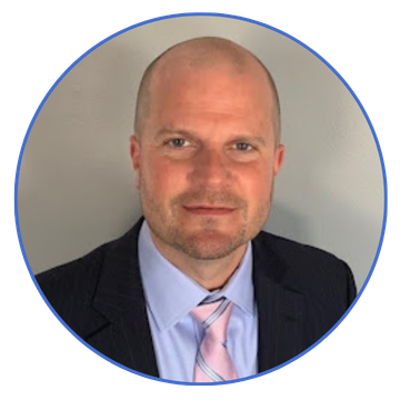 Professional head-and-shoulders portrait of a bald man in a dark suit and light pink tie, looking at the camera, within a circular blue-framed badge.