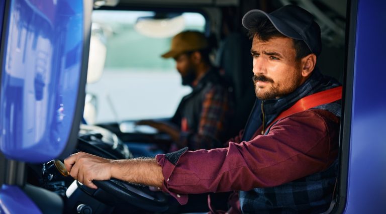 Truck driver in the foreground gripping the steering wheel, wearing a cap and burgundy shirt, inside a blue truck cab with a second driver blurred in the background.