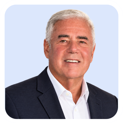 Professional headshot of a smiling older man in a dark suit and white shirt against a light blue background.