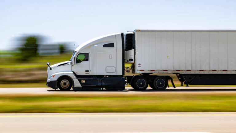 White semi-truck driving at speed on a highway with a blurred background on the left side of the image.